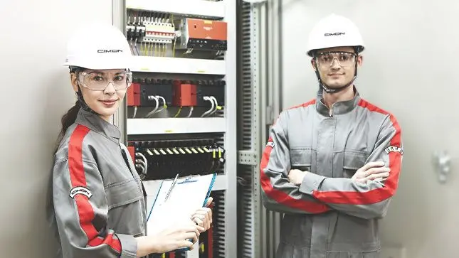 Two workers in CIMON apparel in front of a PLC rack